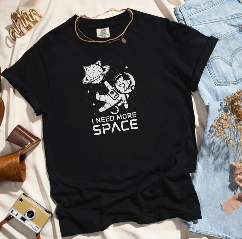 May include: Black t-shirt with a white graphic of a cat wearing an astronaut suit floating in space. The text "I need more space" is printed below the cat.