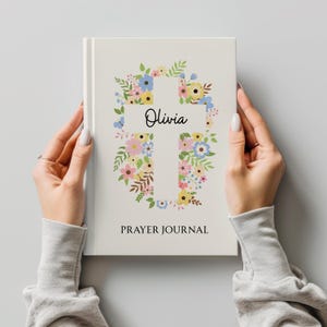 May include: A cream-colored prayer journal with a floral cross design. The cross is filled with colorful flowers and greenery. The name "Olivia" is written in script within the cross, and the words "PRAYER JOURNAL" are printed below.