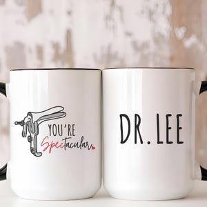 May include: Two white ceramic mugs with black handles. One mug has a black and white illustration of a medical speculum with the text "You're Spectacular" in black and red. The other mug has the text "DR. LEE" in black.