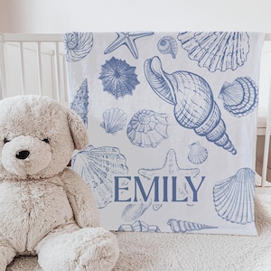 May include: White baby blanket with blue seashell illustrations and the name "EMILY" in blue script. The blanket features various seashell designs, including starfish and conch shells, creating a nautical theme. The blanket is draped over a crib.