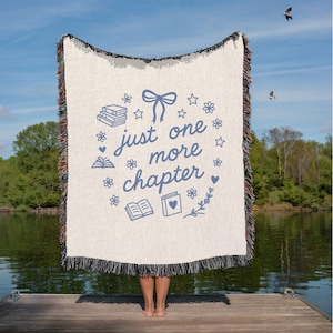 May include: Cream-colored woven throw blanket with the text "just one more chapter" in blue, surrounded by book and floral illustrations. The blanket has a black fringe and is held up against a lake backdrop.