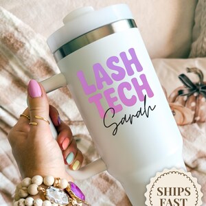 May include: A white tumbler with a handle, featuring the text "LASH TECH Sarah" in purple and pink. The tumbler has a silver rim and a white lid. The image also includes a "SHIPS FAST" sticker.