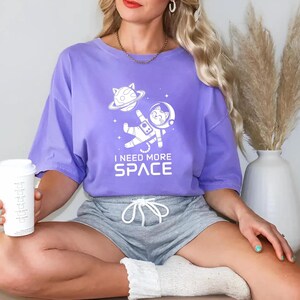 May include: A purple t-shirt with a white graphic of a cat in a spacesuit with the text "I need more space".