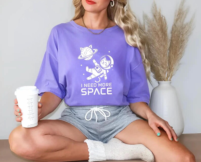 May include: A purple t-shirt with a white graphic of a cat in a spacesuit with the text "I need more space".