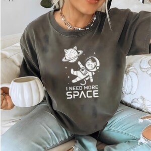 May include: A dark gray t-shirt with a white graphic of a cat in a spacesuit floating in space. The text "I need more space" is printed below the graphic.