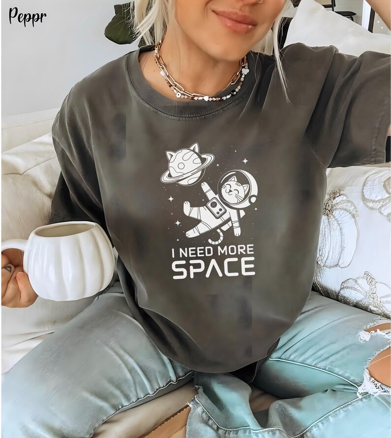 May include: A dark gray t-shirt with a white graphic of a cat in a spacesuit floating in space. The text "I need more space" is printed below the graphic.