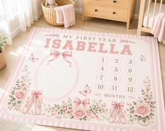Peut inclure: Une couverture de suivi de croissance rectangulaire avec une bordure rayée rose clair et blanche. Le texte indique MY FIRST YEAR ISABELLA. Elle comprend un grand cadre ovale, des chaussons de danse roses, des fleurs, des papillons et les chiffres 1 à 12.