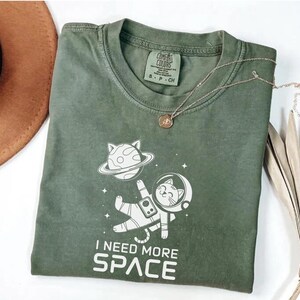 May include: A green t-shirt with a white graphic of a cat in a spacesuit floating in space. The text "I need more space" is printed below the graphic.