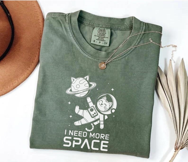 May include: A green t-shirt with a white graphic of a cat in a spacesuit floating in space. The text "I need more space" is printed below the graphic.