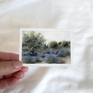 May include: A small watercolor painting of a landscape featuring a tree and blue-purple bushes. The artwork is on textured paper and held by a hand against a white background. The painting has a soft, natural color palette.