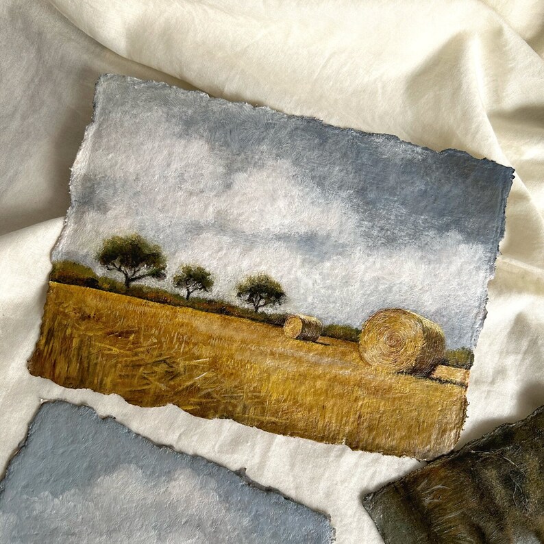 May include: A watercolour painting depicting a field with hay bales. The sky is a light blue with white clouds. There are three trees in the background. The field is a golden yellow colour.