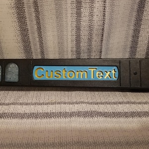 Puede incluir: Un objeto rectangular negro con el texto "Custom Text" en letras amarillas sobre un fondo azul. El objeto tiene una superficie texturizada y está sobre un fondo con estampado gris y blanco.