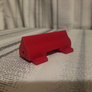 Puede incluir: Un objeto de plástico rojo impreso en 3D con forma rectangular y dos pequeños pies en cada extremo. El objeto tiene un agujero en un lado.