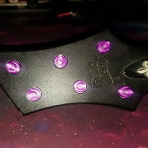 Puede incluir: Guitarra eléctrica negra con tornillos morados y un logotipo de flor de lis. La guitarra tiene el texto "Dean Z  Model" en el cuerpo.