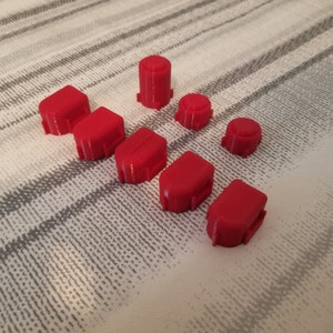 May include: A collection of bright red, 3D-printed plastic components. The assortment includes rectangular and cylindrical shapes, likely parts for a larger product or project. The pieces are arranged on a patterned surface.