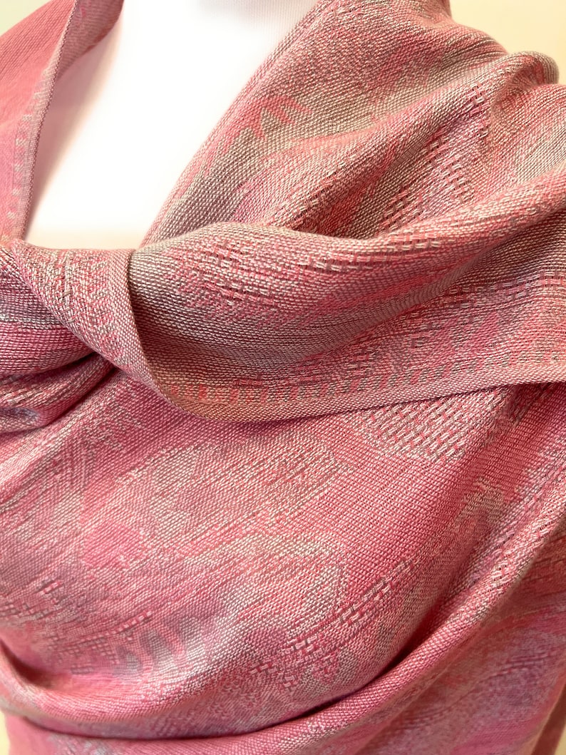 May include: A pink and silver patterned shawl made of a soft, woven fabric. The shawl is draped over a mannequin's shoulders.