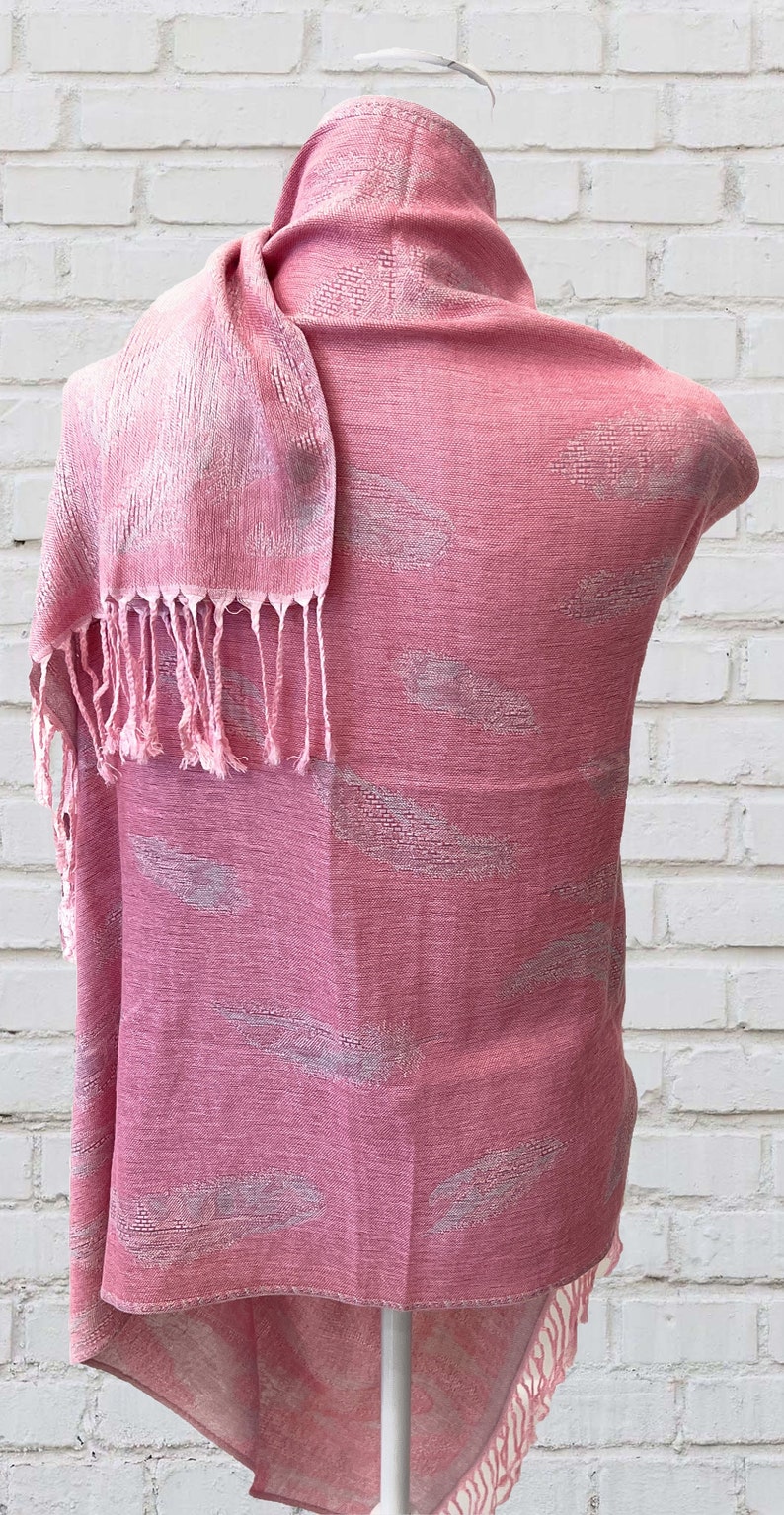 May include: A pink scarf with a white feather pattern. The scarf has a fringe on the bottom and is draped over a white mannequin.