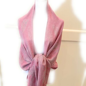 May include: A pink shawl with a subtle shimmer and fringe trim. The shawl is draped over a white mannequin.