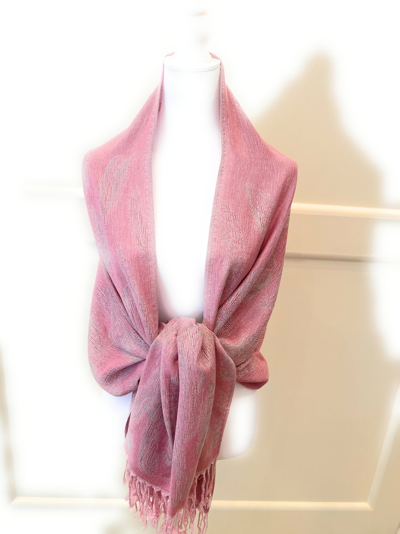 May include: A pink shawl with a subtle shimmer and fringe trim. The shawl is draped over a white mannequin.