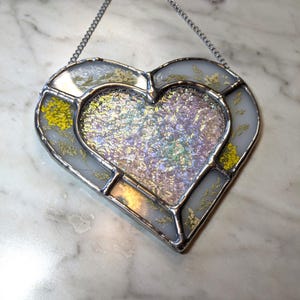 May include: A stained glass heart-shaped ornament with a silver chain for hanging. The heart is filled with iridescent glass and dried flowers in shades of yellow and white.