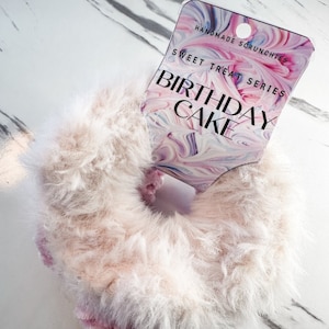 May include: A fluffy pink and white hair scrunchie with a tag that reads "Handmade Scrunchies Sweet Treat Series Birthday Cake".