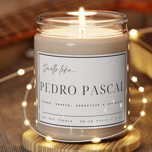 May include: A soy wax candle in a clear glass jar with a white label that reads "Smells like... PEDRO PASCAL FRESH, SMOOTH, SEDUCTIVE & HYPNOTIC SOY WAX CANDLE 50-60 hours / 9 oz". The candle is lit and surrounded by white string lights.