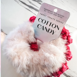 May include: A white and pink fuzzy hair scrunchie with the text "Handmade Scrunchies By: Plush and Petal Sweet Treats Series Cotton Candy" on a white tag.