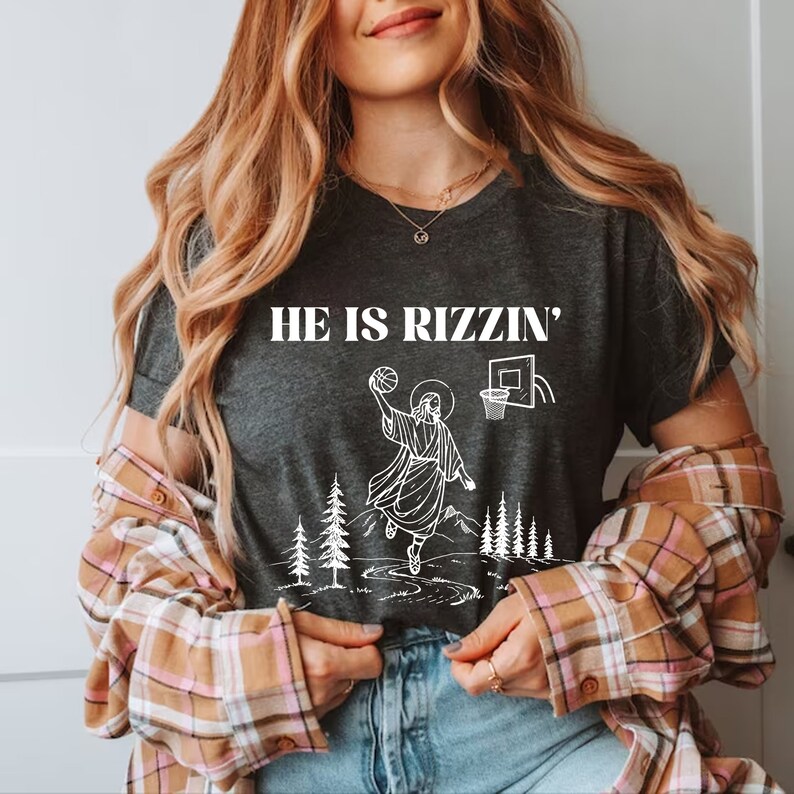 May include: Dark gray t-shirt with the text "HE IS RIZZIN'" above a white graphic of Jesus playing basketball. The graphic includes a basketball, hoop, trees, and mountains.