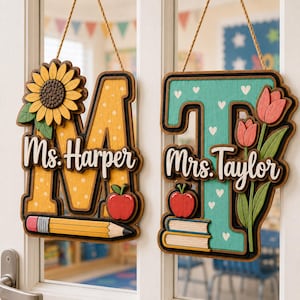 May include: Two hanging wooden letter signs. The left sign is a yellow letter M with a sunflower, a red apple, and a pencil, labeled Ms. Harper. The right sign is a teal letter T with white hearts, tulips, an apple, and books, labeled Mrs. Taylor.