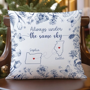 May include: White throw pillow with a blue floral and butterfly design. The pillow features the text "Always under the same sky" with two states and names. A decorative accent for a home.
