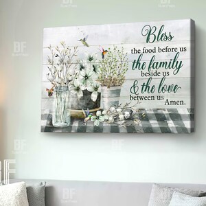 May include: A watercolor painting on a wood background with a white and green checkered tablecloth. The painting features a bouquet of white flowers in a glass jar, a metal bucket, and two hummingbirds. The text reads "Bless the food before us the family beside us & the love between us Amen."