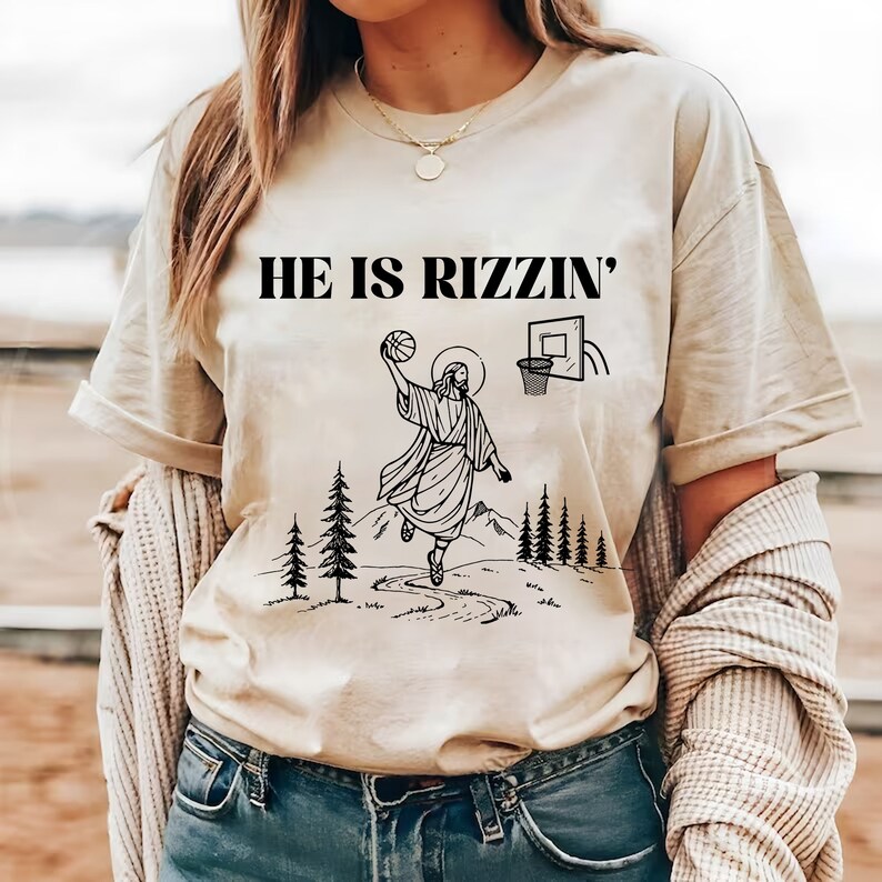 May include: Beige t-shirt with the text "HE IS RIZZIN'" above a black and white graphic of Jesus playing basketball. The graphic includes a basketball hoop, trees, and a path.