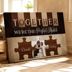 May include: A brown and beige wood-grain canvas print with the text "Together We're the Perfect Match" and two puzzle pieces with the names "Donald" and "Ashley" on them. A photo of a couple is in the center of the puzzle pieces.