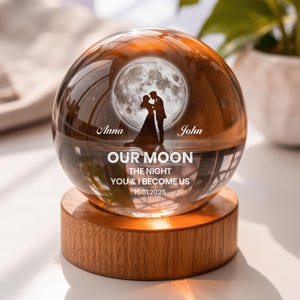 May include: A clear glass sphere resting on a cylindrical wooden base. Inside the sphere is a laser-etched image of a couple standing before a full moon, with the text Anna, John, OUR MOON, THE NIGHT YOU & I BECOME US, and 15.01.2025.