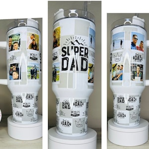 May include: White stainless steel travel mug with a black and white design featuring the words "Super Dad" and a crown. The mug has a black handle and a black lid. The mug is surrounded by a white background.