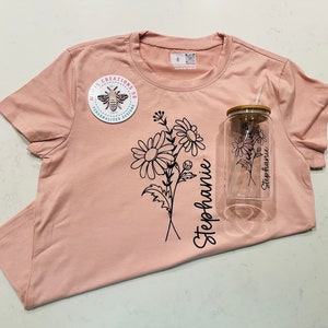 Könnte beinhalten: Ein pinkfarbenes T-Shirt mit einem schwarzen Blumenmuster und dem Namen "Stephanie" darauf gedruckt. Eine durchsichtige Glaswasserflasche mit einem Holzdeckel und einem Strohhalm ist ebenfalls abgebildet. Das T-Shirt hat einen kleinen Bienensticker mit dem Text "M.I.S Creations SD Personalized Designs".