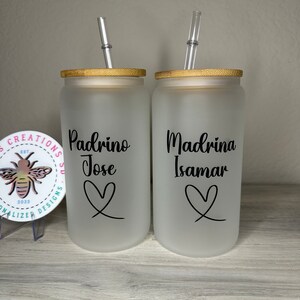 May include: Two frosted glass tumblers with bamboo lids and clear straws. The tumblers have black lettering that reads "Padrino Jose" and "Madrina Isamar" with a heart design below each name.