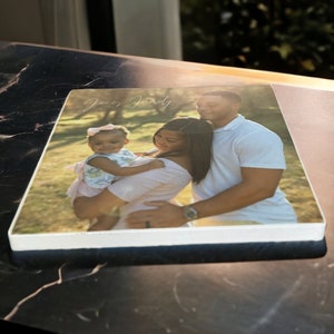 May include: A photograph of a family of three. A woman is holding a young girl in her arms. The woman is wearing a pink shirt and the girl is wearing a light blue and white dress. A man is standing behind them with his arm around the woman. He is wearing a white shirt. The photo is printed on a white square tile with a light brown background.