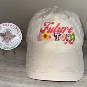Könnte beinhalten: Eine beige Baseballkappe mit einem weißen Schirm und dem Text "Future Doctor" in bunten Buchstaben mit einem Blumendesign.
