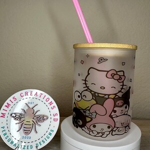 Op de afbeelding: Een frosted glazen beker met een bamboe deksel en een roze rietje. De beker heeft een kleurrijke afbeelding van Hello Kitty en andere cartoonfiguren. De beker staat op een wit voetstuk. Een ronde sticker met een bijenontwerp en de tekst "Mimi's Creations SD Est. 2023 Personalized Designs" is op de voorgrond.