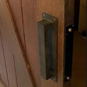 May include: Close-up of a wooden door with a dark metal handle and a black metal plate. The door is made of stained wood with diagonal paneling. The handle is rectangular and mounted on a rectangular plate.