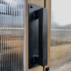 May include: A black, rectangular door handle with a square grip is mounted on a light-colored wooden door. The door features a textured glass panel and a black latch.