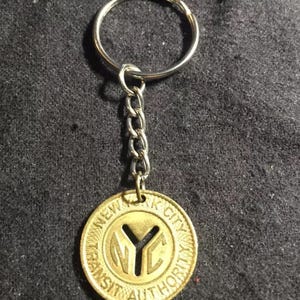 May include: A gold-colored keychain featuring a replica of a New York City Transit Authority token. The token has the words "New York City Transit Authority" around the edge and a stylized "NYC" in the center.