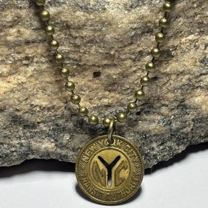 May include: A brass-toned necklace with a circular pendant displaying the text "NEW YORK CITY TRANSIT AUTHORITY" and a "Y" symbol. The necklace has a ball chain, and the pendant hangs from the chain.