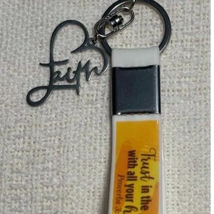 May include: A silver-toned keychain with a rectangular charm featuring the text "Trust in the Lord with all your heart." A heart-shaped charm with the word "Faith" is also attached. The keychain has a silver clasp and ring.