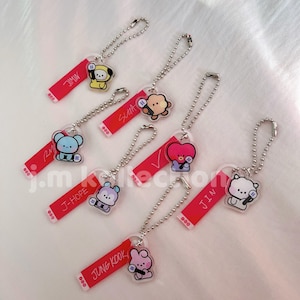 May include: Seven acrylic keychains featuring cartoon characters and red rectangular tags with names printed in white. Each keychain includes a silver ball chain. The names include Jimin, Suga, RM, V, J-Hope, Jin, and Jung Kook, each paired with a unique character.