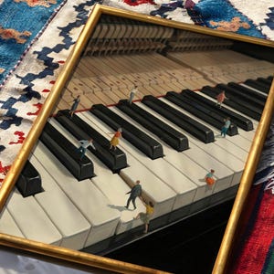 May include: A framed artwork depicts a piano keyboard with miniature figures interacting on the keys. The gold frame surrounds the image, which features black and white keys and a detailed interior. The background includes a colorful rug.