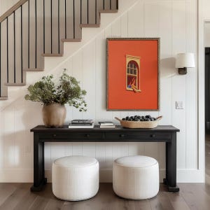 May include: A black console table with two drawers, topped with books and a woven basket of dark objects. Two round, upholstered ottomans sit in front of the table. A framed artwork with an orange background hangs on the white paneled wall.