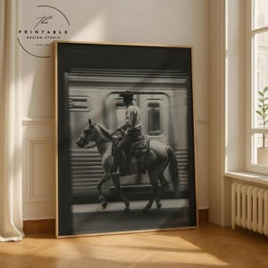 Könnte beinhalten: Schwarz-Weiß-Druck mit einem Cowboy auf einem Pferd vor einem Zug. Das Bild hat eine Vintage-Ästhetik. Der Druck wird in einem Raum mit Fenster und Heizkörper ausgestellt.