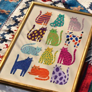Puede incluir: Una impresi&oacute;n art&iacute;stica enmarcada con ilustraciones de gatos coloridos y estampados. Cada gato presenta patrones &uacute;nicos, como rayas y lunares, en varios colores. La obra de arte est&aacute; enmarcada en un marco dorado y sobre un fondo crema. Los gatos est&aacute;n en varias poses.